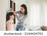 Happy Asian mother using book and pencil to measure height of little girl near wall at home