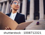 Thoughtful African American businesswoman in optical spectacles holding documents looking away while standing in urban setting with copy space. Female dark skinned student of high economic university