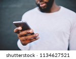 Cropped image of african american man holding modern smartphone dialing number, dark skinned male in white shirt using mobile phone for chatting in social networks via 4G internet connection outdoors