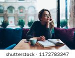 Dreaming beautiful hipster girl looking away while resting at cafeteria and writing romantic essay, attractive young woman in eyeglasses enjoying free time and planning to do list in notebook