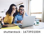 Small photo of Cheerful man and woman with pencil in had near face smiling while planning further work together and using laptop at home