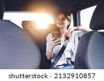 Cheerful positive young female in casual wear sitting in automobile backseat with seatbelt fastened and looking away while using mobile phone