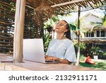 Joyful graphic designer in eyeglasses using netbook computer during remote working outdoors, cheerful female copywriter with laptop laughing and rejoicing - digital nomad and freelance lifestyle