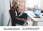 Small photo of Smiling senior caucasian mature businesswoman sitting on chair near table and talking on smartphone. Concept of small business and entrepreneurship. Modern successful woman. Home art studio. Side view