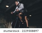 Low angle of young male in headphones looking away while sitting on spinning stationary cycle and listening to music in wireless headphones while doing cardio workout in gym