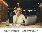 Positive young female freelancer sitting with hand touching cheek at wooden table with smartphone diary pen juice glass and looking at screen of laptop while working on remote project in cafe
