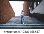 African American female in casual clothes walking downstairs near brick wall with paper coffee cup in hands and looking at camera in city street on weekend