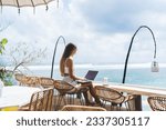 Side view full body of young barefoot female freelancer with long hair sitting at table on terrace and working remotely via laptop during vacation in Bali