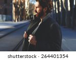 Portrait of fashionable well dressed man with beard posing outdoors looking away, confident and focused mature man in coat standing outside at sunny evening, elegant fashion model