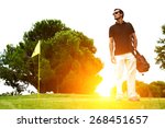 Full length portrait of stylish golfer in glasses standing on golf course with amazing flare sunset light on background, handsome man with golf bag on shoulder standing on golf course, filtered image