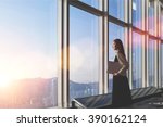 Successful female office worker with net-book is standing in skyscraper interior against big window with city view on background. Proud asian woman architect looking satisfied with completed project