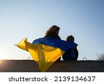 little boy and a woman sit with their backs at sunset against the sky with a blue and yellow Ukrainian flag. Family, refugees, unity, support. Ukrainians are against the war. stand with Ukraine