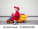 Small photo of cute smiling 3-4 year old boy in red overalls uniform and cap is sitting on big toy car truck with cardboard boxes parcels. postman's little helper, truck driver. Business delivery advertising