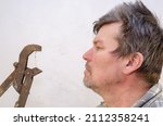 Small photo of Portrait of a geezer talking with a dusty pipe wrench. Profile of an Unshaven Man and a Rusty Hand Tool. Adult male with ruffled hair. Indoors. Selective focus.