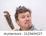 Small photo of Portrait of a funny geezer with a rusty old dirty pipe wrench. An unshaven man with a hand tool covered in cobwebs. Adult male with ruffled hair. Indoors. Selective focus.