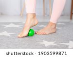 close-up two feet with a pedicure of a young woman, one foot stands on a green spiky massage ball on a carpet in a home interior.
