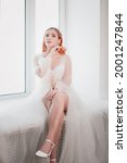Small photo of a full-length young bride with red hair in a white transparent cape or negligee is sitting on the window in the morning.