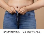 Small photo of Caucasian female with big belly and overweighted sides trying to zip up blue jeans. Visceral fat. Body positive. Sudden weight gain. Tight little clothes. Need for wardrobe change.