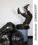 Small photo of Businessman in Suit Stuck Upside Down in Trash Can Next to Garbage Bag Pile. Concept of Over a Barrel. Thrown Away by Capitalism and Greed.