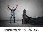 Small photo of A happy businessman with boxing gloves on arms raised in victory stands near a giant male leg fallen down. Fight off competition. Unexpected winner. Chance of success.