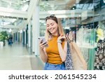 Young woman consumer in the mall browses chat and uses using a smartphone. female standing with a mobile phone in her hands in shopping center. indoor. happy shopper girl with gift bags make purchases
