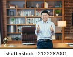 Small photo of asian boss holding thumbs up on hands looking at camera and smiling, man working in office, portrait of successful and affirmative businessman