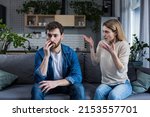 Family quarrel, man and woman sitting on sofa at home. angry woman yells at her husband, blames. The man is silent. He listens, holds his head, embarrassed