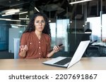 Small photo of Frustrated and sad business woman with phone in hands looking at camera, latin american woman working with laptop inside modern office building, unclear emotional state of female employee