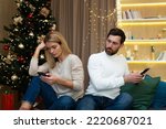 Family couple quarreled on Christmas sitting at home on sofa, husband and wife not talking to each other, holding phones in hands browsing social networks, near Christmas tree.