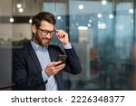 Small photo of Successful financier investor works inside office at work, businessman in business suit uses telephone near window, man smiles and reads good news online from smartphone.
