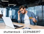 Small photo of Cheated and disappointed businessman working inside office, man holding bank credit card and trying to make a purchase in online store, online fraud and deception.