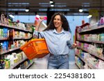 Small photo of Portrait of a happy woman shopper in a supermarket, a Hispanic woman with a basket of goods smiles with pleasure and dances among the shelves with goods, a satisfied shop customer.