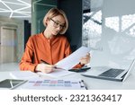 Small photo of Serious and pensive business woman behind paper work inside office, female financier worker thinks about contracts and reports with charts and graphs, blonde successful woman uses laptop at work.