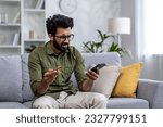 Small photo of Upset man at home cheated and denied money transfer, hispanic sitting on sofa at home with phone and bank credit card, cheated and disappointed in living room.