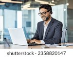 Young successful man at workplace with headset phone typing on laptop keyboard, customer support worker for online buyers inside office, hispanic helping and consulting customers.