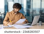 Young serious focused and thinking businessman paper work, successful hispanic man reviewing contract reports and invoices, sitting at desk inside office with laptop, checking and filling form.