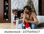 Bored and sad man sitting on sofa for christmas near christmas tree, hispanic depressed in living room lonely no friends for new year celebration.
