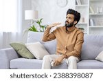 Small photo of Young Indian man with beard sits in the living room in summer, fanning his hand, suffering from the heat, holding the air conditioner remote control, unpleasant temperature in the room, overheating.