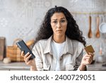 Small photo of Sad girl holding phone and card while sitting in kitchen at home, failed online payment, failed shopping, scams on the internet.