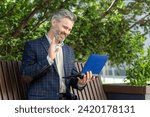 Small photo of Affable senior businessman with a tablet engaging in a video call waves hello from a bench outside an office building.