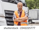 Small photo of Experienced road haulier professional stands in orange vest in front of the vehicle with arms crossed.