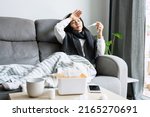 Small photo of Sick Asian woman feeling cold sitting on sofa at home. Girl has a high fever sit on couch with tissue, medicine, a cup of warm water on desk. Health problems, allergy, season change fever concept.