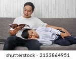 Small photo of Asian young fathers in white tshirt reading a book while his son in white sweater fast asleep