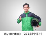 Portrait of Asian online taxi driver wearing green jacket showing thumb up hand gesture and holding helmet. Isolated image on white background