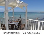 Small photo of SAN TEODORO, SICILY - 06 15 2022: Wooden table with plates of food and people swimming in the sea at the Plaia Del Rio Marsala beach in the Sicilian village of San Teodoro