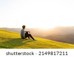 Young man sitting on the grass and watching the sunset in the mountains in the background. Free and happy person enjoying the sunset