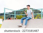 Happy man sitting on top of a skate ramp, with a longboard on his side, while using his cell phone or smartphone to chat online or access social networks.
