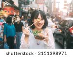 Poeple travel and eating street food concept. Happy young adult asian foodie woman holding spicy grilled squid at southeast asia Chinatown market.