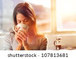 Woman drinking coffee at home with sunrise streaming in through window and creating flare into the lens.