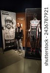Small photo of LeMans, France; October 9, 2023: Young blonde girl with long hair with a photo camera on vacation at the 24h LeMans museum next to the tribute poster of Fernando Alonso and his fireproof monkey